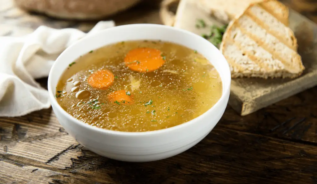 Steaming cup of homemade chicken stock with herbs and vegetables in the background